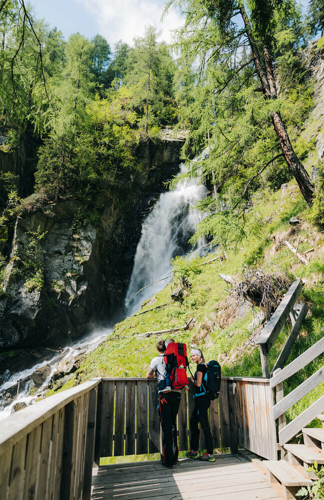 Eine junge Familie steht auf einer hölzernen Plattform und sieht auf den Wasserfall Kristeinertal.