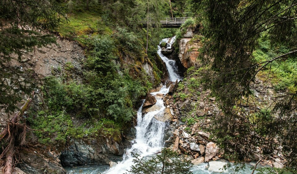 Ein kleiner Wasserfall schlängelt sich durch einen Wald. Im Hintergrund befindet sich eine kleine Holzbrücke, die darüber führt.