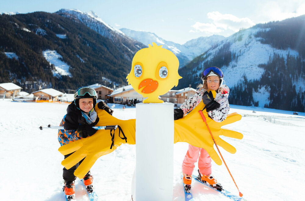 Zwei Kinder stehen im familienfreundlichen Skigebiet Golzentipp in Obertilliach hinter einem gelben Kurvenzeiger in Entendesign mit winterlichem Bergpanorama im Hintergrund.
