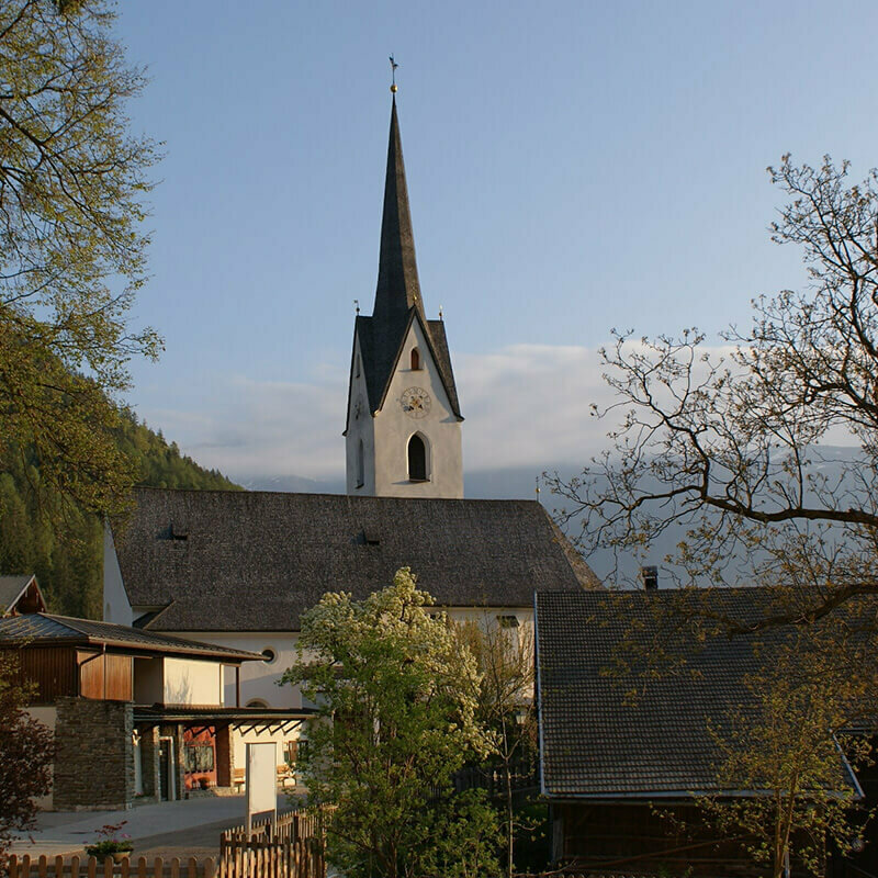 Die Kirche in Leisach inmitten des Ortskerns mit herbstlichen Bäumen und Hochnebel über dem Zettersfeld im Hintergrund.