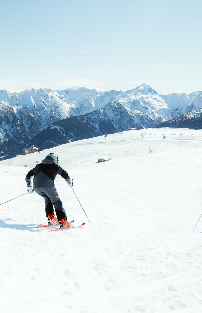 Zwei Skifahrerinnen auf einer flachen Piste oberhalb der Baumgrenze. Im Hintergrund der Taleinschnitt und ein verschneiter Bergkamm, der den Abschluss zur milchig wirkenden Bewölkung an einem Schönwettertag im Winter zeigt.