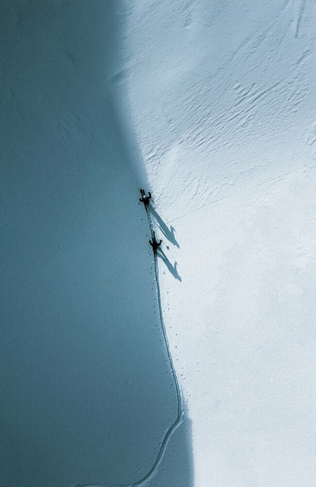 Zwei Skitourengeher gehen auf ihren Skiern entlang einer Schattenlinie, die der Berg auf die frisch verschneite Landschaft wirft.