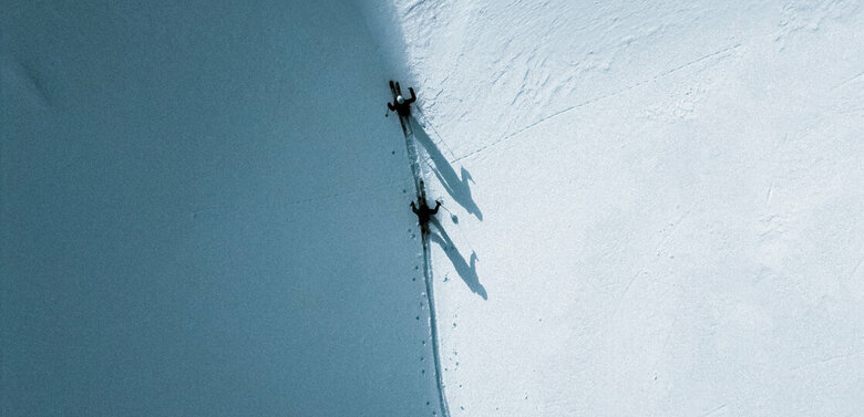 Zwei Skitourengeher gehen auf ihren Skiern entlang einer Schattenlinie, die der Berg auf die frisch verschneite Landschaft wirft.