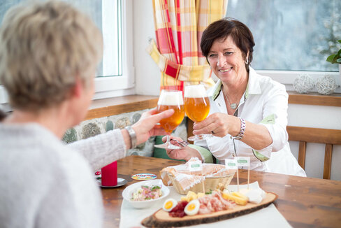 Zwei Frauen stoßen bei einer Brettljause mit verschiedenen Käsesorten in Würfel geschnitten, feinen Speck- und Henkelescheiben und hartgekochten Eiern mit einem Glas Bier an.