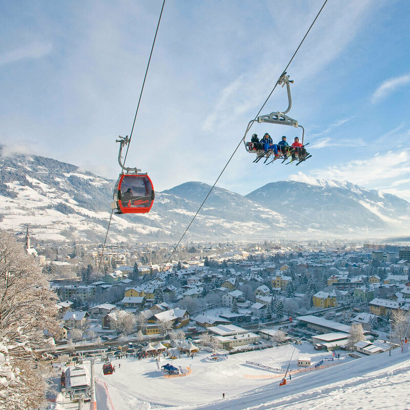 Hochstein Lienz Sessel und rote Gondel schweben über die tief verschneite Winterlandschaft am Hochstein mit der Stadt Lienz im Hintergrund.