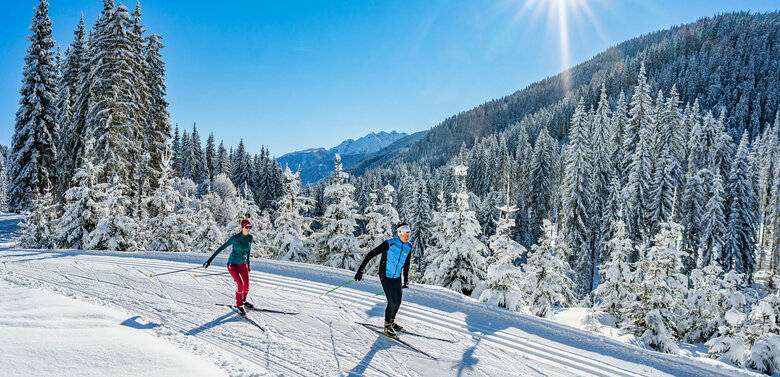 Zwei Langläufer:innen laufen bei strahlendem Sonnenschein auf der Loipe in Obertilliach mit einem frisch verschneiten Wald im Hintergrund