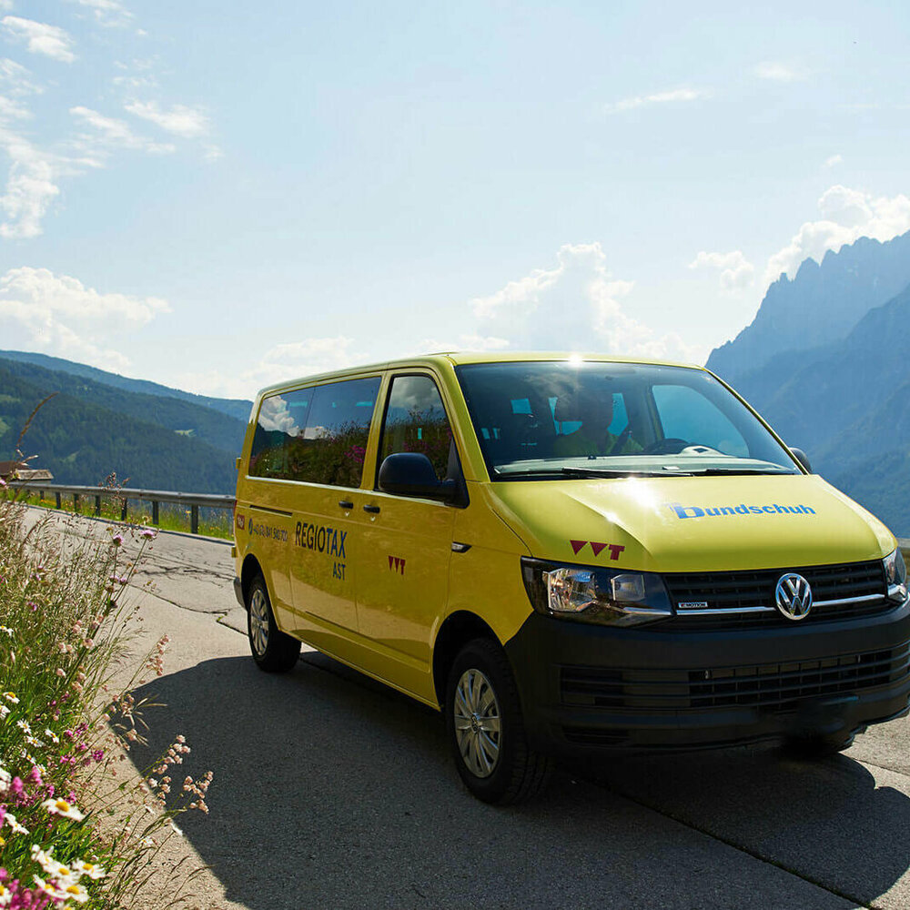 Das gelbe Autosammeltaxi Osttirol führt über eine Straße, daneben befindet sich eine Blumenwiese. Im Hintergrund sind Berge.