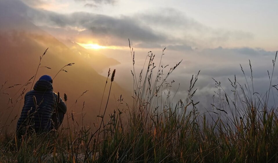 Eine Person mit dunkler Jacke und Mütze sitzt im hohen Gras und blick auf die untergehende Sonne in der Ferne. Im Tal hängt eine dicke Wolkendecke.