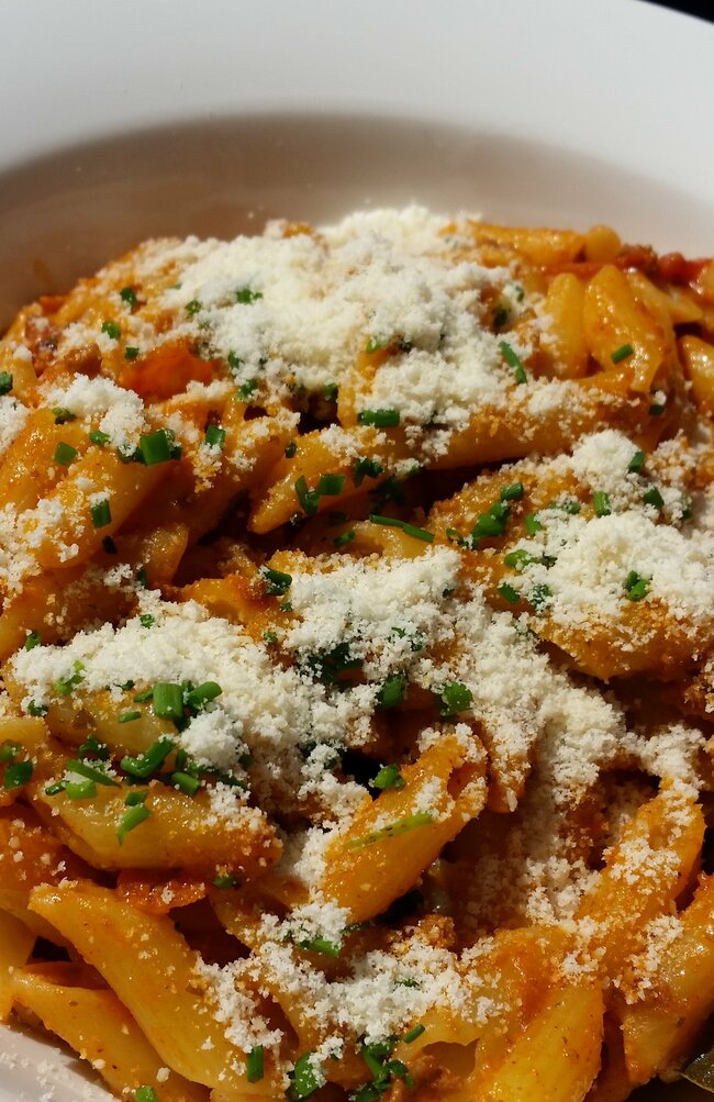 Weißer, runder Teller auf Holztisch mit Penne-Nudeln mit pikanter Sauce und darübergestreuter Parmesan