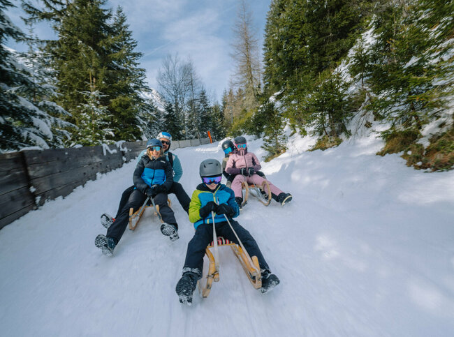 Eine Familie in Skianzügen rodelt auf ihren Schlitten talwärts auf der Rodelbahn im Winkeltal in Außervillgraten, Osttirol.