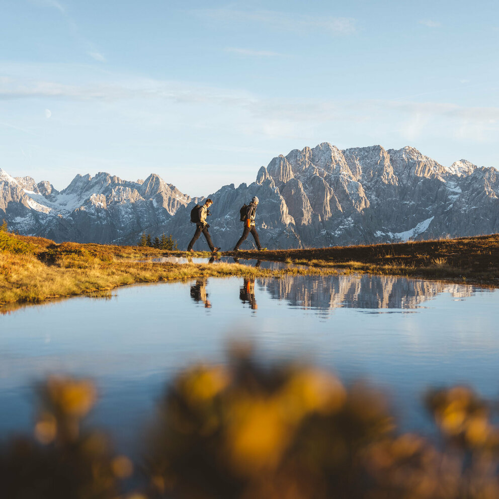 Der See am Hochstein spiegelt zwei vorbei Wandernde und die Bergkulisse in seinem klaren Wasser.