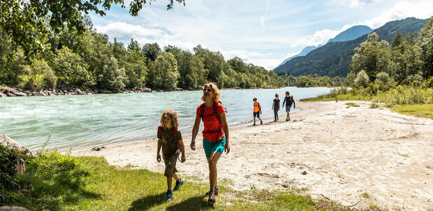 Mehrere Personen in Sportbekleidung spazieren am Ufer der Isel entlang des Iseltrails an einem sonnigen Tag.