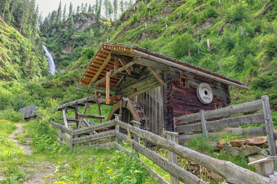 Ein Bild der "Stadtner Mühle" in Maria Hilf, im Hintergrund Blick auf den tosenden Wasserfall des Stallebaches. Das dunkle Holz der Mühle hebt sich deutlich von der grünen Umgebung des umliegenden Waldes ab.