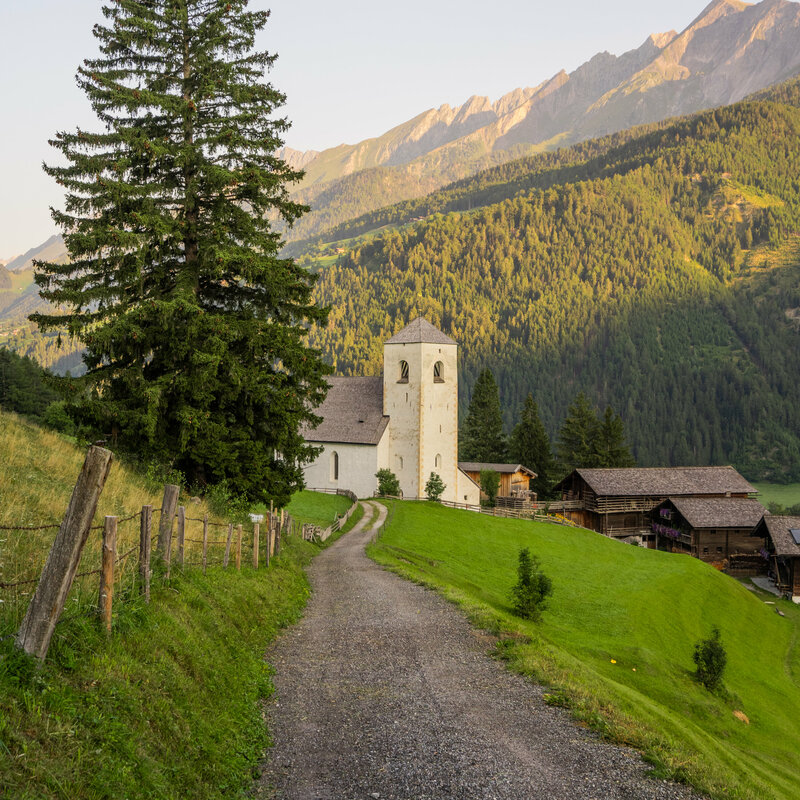 Ein dunkelgrauer Schotterweg führt vorbei an einem großen dunkelgrünen Tannenbaum in Richtung der weißen St. Nikolauskirche. Neben der Kirche stehen drei alte Bauernhäuser, die aus dunkelbraunen Holz errichtet wurden - auf einem der drei Bauernhäusern sieht man auf einem Balkon violette Blumen. Im Hintergrund der Bauernhäuser tauch das warme Abendsonnenlicht die dunkel grünen Wälder und sandfarbenen Berge in ein leicht orangefarbenes Licht.