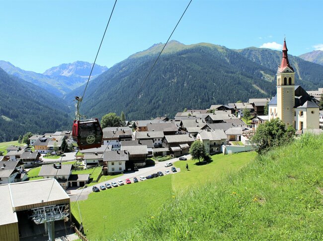 Das Dorf Obertilliach im Tiroler Gailtal, rechts im Bild die markante Kirche, links die bergwärts fahrende Gondelbahn vor alpiner Kulisse