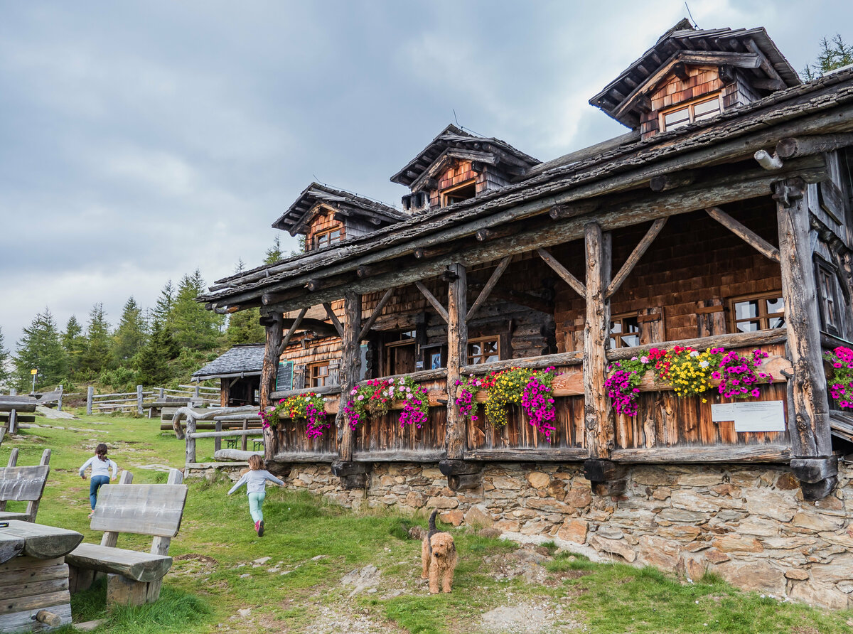 Anna Schutzhaus Das urige Anna Schutzhaus aus Holz mit Blumendeko und spielenden Kindern und einem Hund davor.