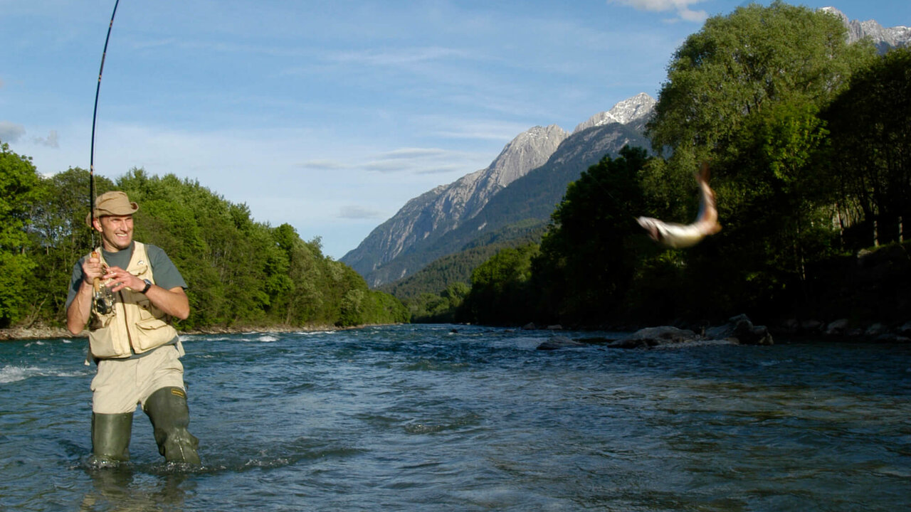Ein Mann mit Hut steht in einem Fluss und ist am Fliegen fischen. An seiner Angel schwingt ein Fisch.