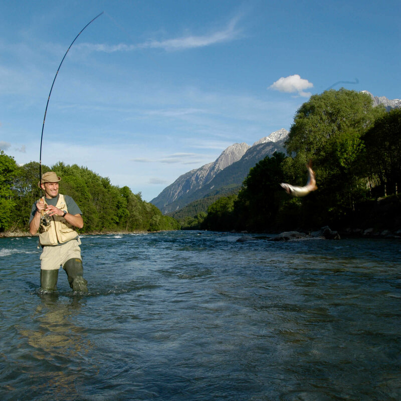 Fliegen fischen Ein Mann mit Hut steht in einem Fluss und ist am Fliegen fischen. An seiner Angel schwingt ein Fisch.