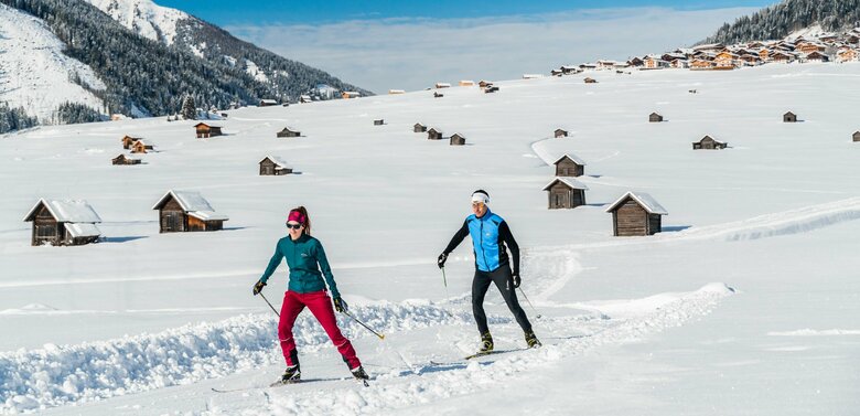 Zwei Wintersportler laufen auf ihren Langlaufskiern auf der Loipe in den Obertilliacher Feldern mit den zahlreichen kleinen Hütten und dem Dorf im Hintergrund