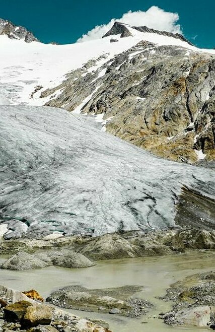 Eine nach oben spitz zulaufende Pyramide, auf welcher Iseltrail oben steht, steht vor einer gewaltigen Bergkulisse des Umbalkees. Der Schnee ragt noch fast bis zur Pyramide, umgeben vom grauen Gestein der Bergwelt und des strahlenden Blau des Himmels.