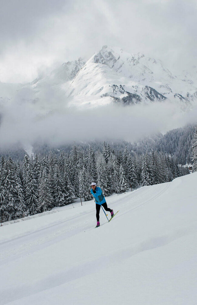 Langlaufen Obertilliach Zwei Langläufer:in auf der Loipe mit einem frisch verschneiten Wald und einem winterliche Bergpanorama im Hintergrund