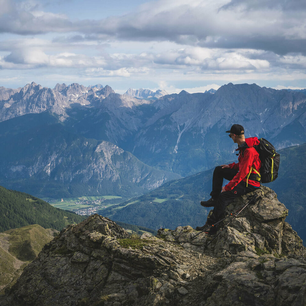 Ein Wanderer mit roter Jacke, schwarzem Rucksack und Kappe sitzt auf einem Felsbrocken in der Schobergruppe und blickt ins Tal Richtung Oberlienz.