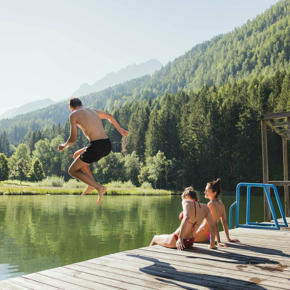baden-tristacher-see Zwei Mädchen sitzen auf einem Holzsteg und ein Junge springt ins Wasser am Tristacher See.