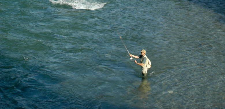 Fliegen fischen Blick von oben auf einen Mann im Fluss mit einer Angel beim Fliegen fischen. Am Ufer wachsen saftig grüne Bäume und im Hintergrund ragen Berggipfel in die Höhe.