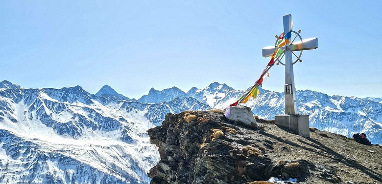 Gipfelkreuz am Figerhorn im Frühling mit noch winterlichen Berggipfeln im Hintergrund.